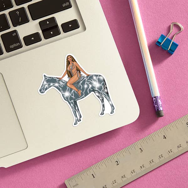 A sticker of a woman with a medium-dark skin tone and long hair sitting on a silver horse and looking at the viewer. The sticker is attached to a Macbook.