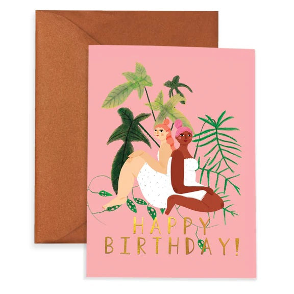 A pink greeting card with an illustration of two women sitting back to back before plants. They have a light and dark skin tone and are wearing white dresses. The card reads "Happy Birthday!"