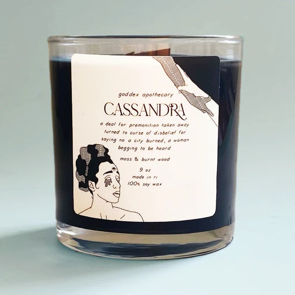 A dark candle in a glass jar. A sticker with an illustration of a woman's face and text is printed on a sticker on the jar.