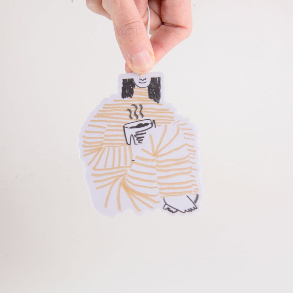 An air freshener of a woman sitting, holding a cup of tea. She is wearing an orange striped pajama.