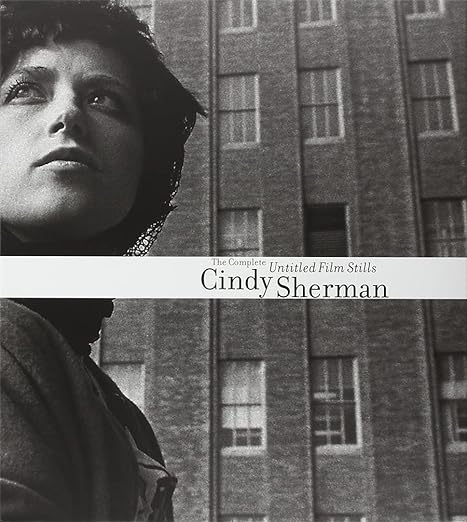 A book cover with a black and white photograph of a woman before a building. The title reads "Cindy Sherman: The Complete Untitled Film Stills."