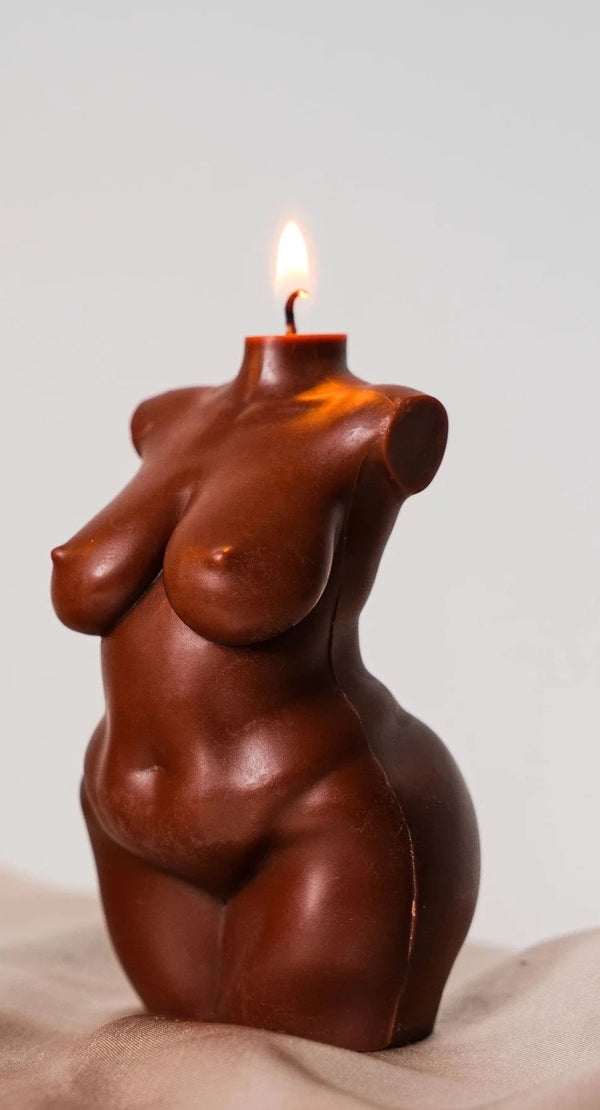 A white background with a candle in the foreground. The candle is in the shape of a nude woman's body. The woman has a dark skin-tone. The wick, which is above the woman's neck, is lit with a flame. 