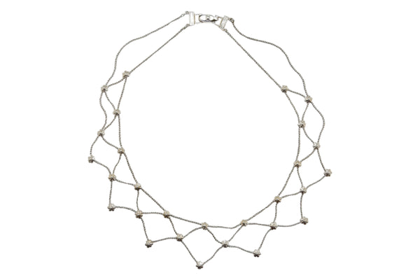 A white background with a silver necklace before it. The necklace is made up of a lattice of silver chains with floral accents. 