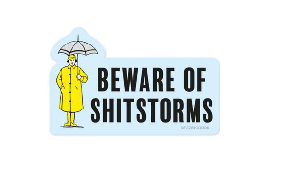 A white background with a sticker in front of it. The sticker is blue and features a woman in a yellow raincoat. She is holding a grey umbrella. There are words on the sticker that say "Beware of Shitstorms."