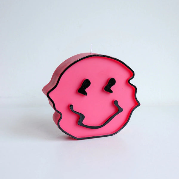 A white background. Before it is a pink, wavy candle in the shape of a smiley face. The smiley face is outlined in black wax and has black features for the eyes and mouth.