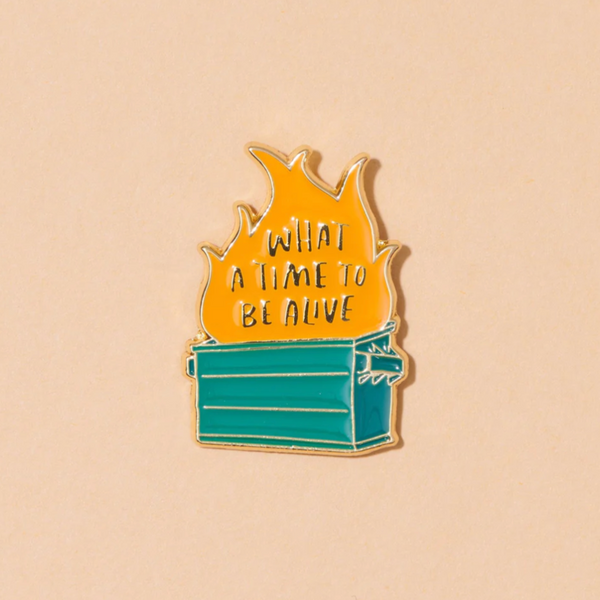 A peachy background with a pin before it. The pin is an illustration of a dumpster fire with the words "What a Time to be Alive" written in the flames. 