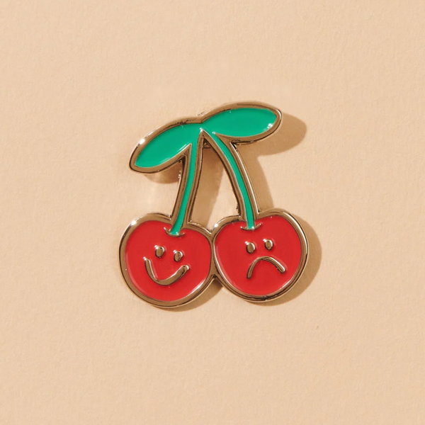 A peachy background with an enamel pin before it. The pin is in the shape of two cherries, one with a smiling face and one with a sad face. 