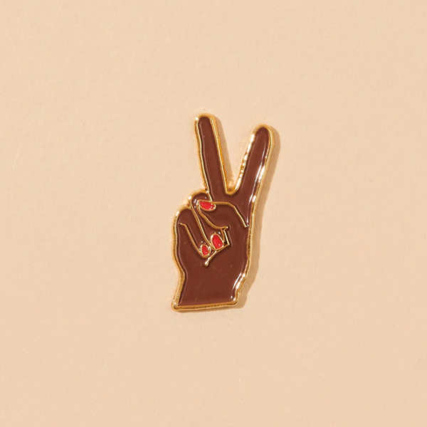 A peachy background with an enamel pin before it. The pin is a peace hand with a dark skin-tone and a red manicure holding up a peace sign. 