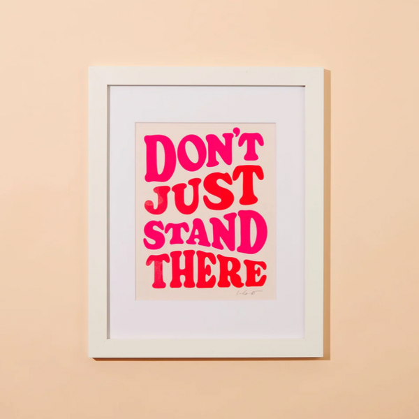 A peachy background with a framed print before it. The print has the words "Don't Just Stand There" in neon pink print. 