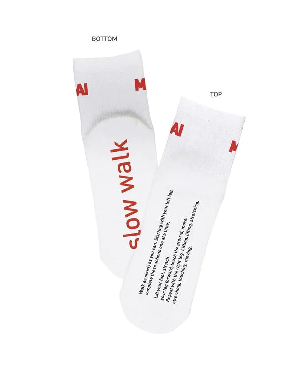 A white background with white socks before it. The socks feature the words "Slow Walk" on the bottom. Below it are instructions on how to walk in a way that is mindful. On the top of the socks are the letters "MAI" in red lettering. 