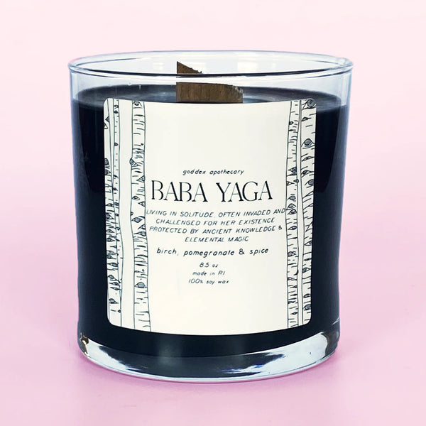 A black candle in a glass before a pink background. The text on the candle reads "Baba Yaga, Birch & Pomegranate Soy Candle."