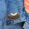 A golden and black enamel pin of a collar with the word "Dissent" written on top of it, attached to a denim jacket.