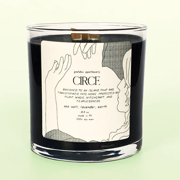 A black candle in a white glass with a sticker attached to it, reading "Circe. Sea Salt, Lavender & Earth Soy Candle" and an illustration of a woman.