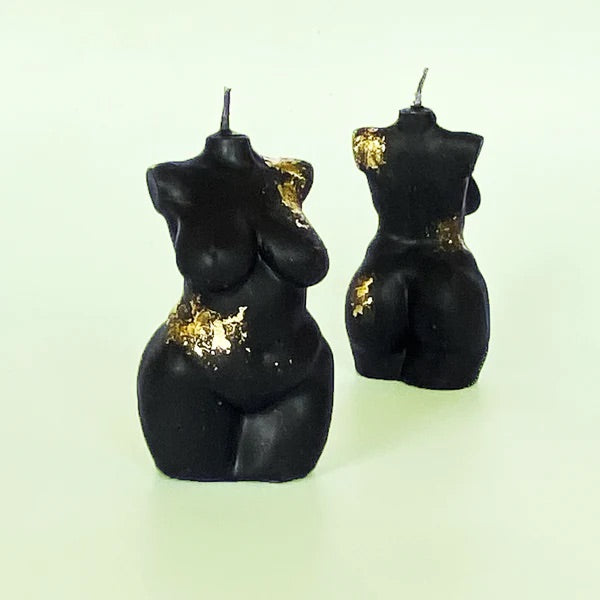 Two black candles in the shape of female torsos. There are some golden highlights on the candles, adding a glamorous element.