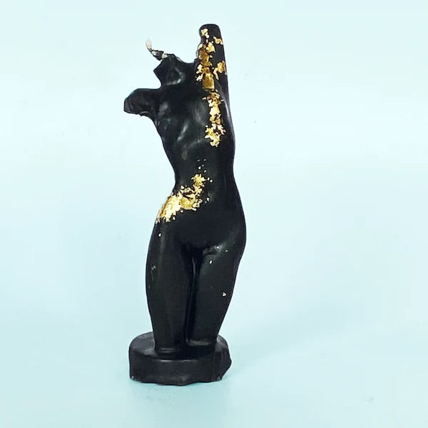 A black candle in the shape of a female torso. Little golden highlights are added onto the body.