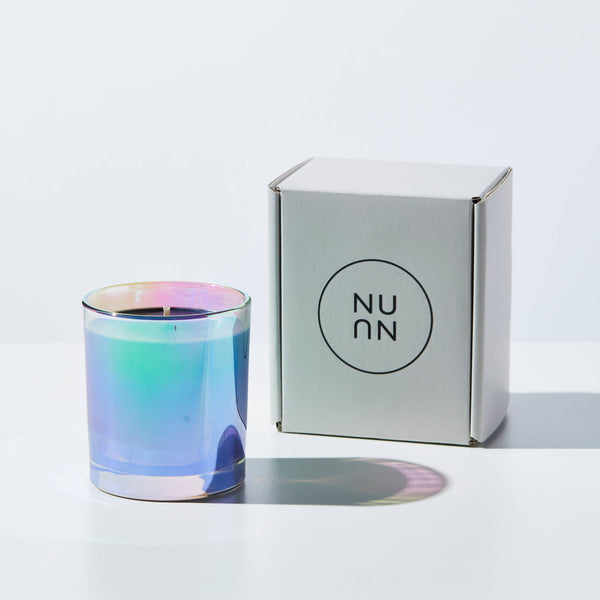 A candle standing next to a paper box. The candle is in shiny and colorful glass jar, reflecting light.
