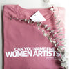 A folded pink t-shirt is lying on a black background. On the t-shirt, text in white capital letters reads: "Can you name five women artists?"