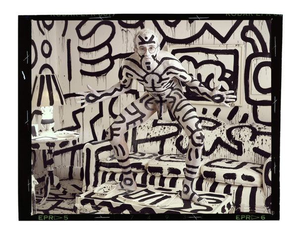 Photograph of a man painted in geometric forms standing in a room painted in the same pattern.