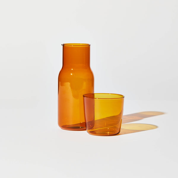 A carafe and cup set in amber standing on white surface casting a yellow shadow.