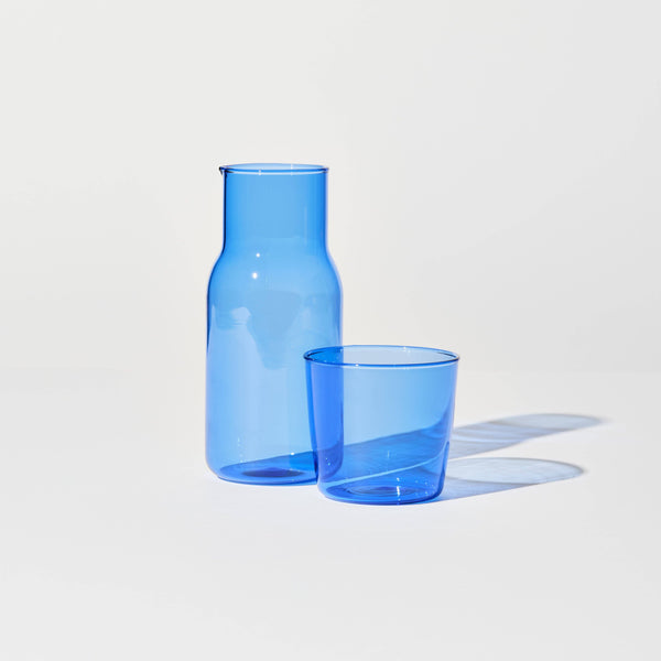 A carafe and cup set in blue standing on white surface casting a blue shadow.