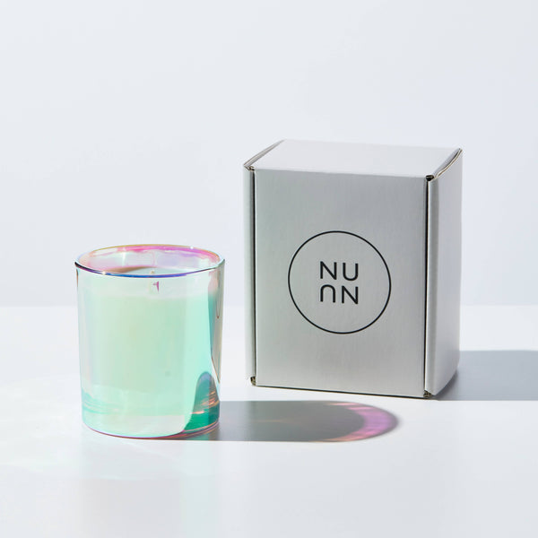 A candle in a shimmering glass standing next to packaging in front of a white wall. On the packaging, it reads "Nu" written twice in a circle.