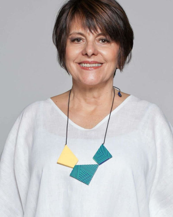 A woman wears a necklace and smiles at the camera. The necklace features teal and blue stripped geometric beads accented with yellow, and hangs on an adjustable black cord.