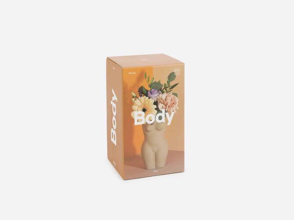 A box showing a vase in the shape of a female torso and legs in beige clay. Several light colored flowers are in the vase. 