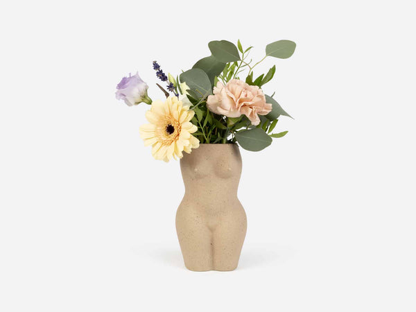 A vase in the shape of a female torso and legs in beige clay. Several light colored flowers are in the vase. 