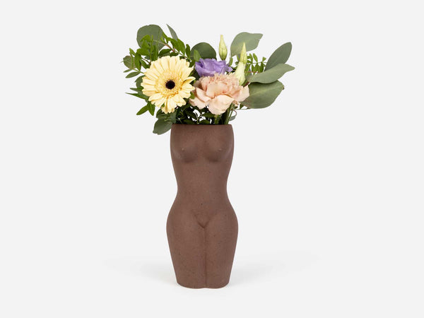 A vase in the shape of a female torso and legs in brown clay. Several light colored flowers are in the vase. 