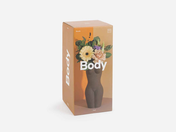 A box showing a vase in the shape of a female torso and legs in brown clay. Several light colored flowers are in the vase. 