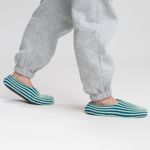 A person with gray sweatpants is wearing indoor knit slippers with chunky rib stripes in teal and light blue.