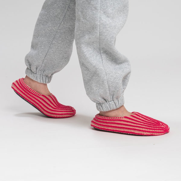 A person with gray sweatpants is wearing indoor knit slippers with chunky rib stripes in peach and pink.