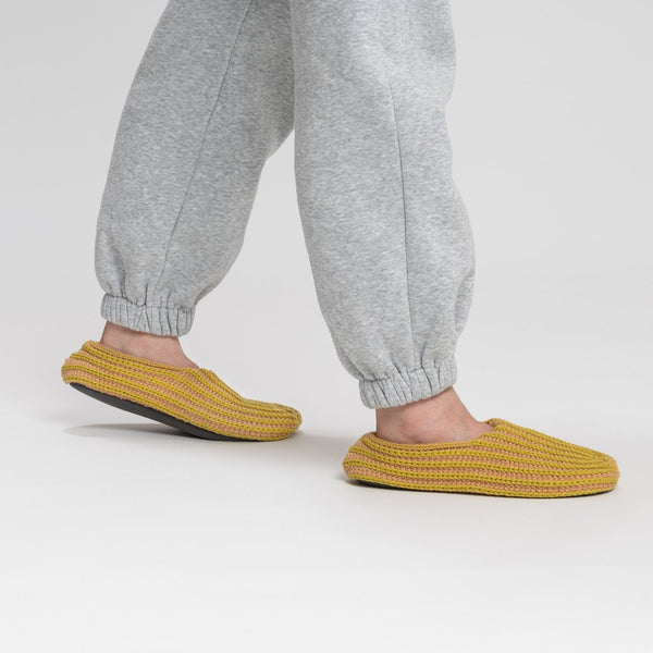 A person with gray sweatpants is wearing indoor knit slippers with chunky rib stripes in mustard yellow and beige.