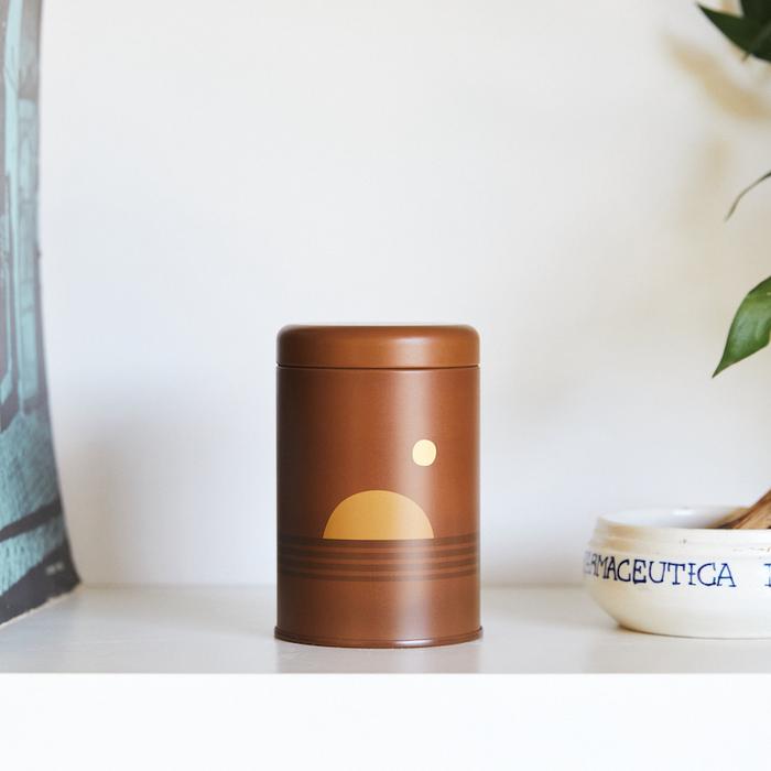 Candle in a brown metal can with an illustration of a sunset. The candle is standing next to a plant.