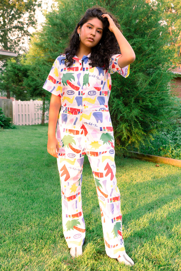 A woman with a medium skin tone wearing a white pair of overalls with a colorful abstract print in red, blue, and yellow.