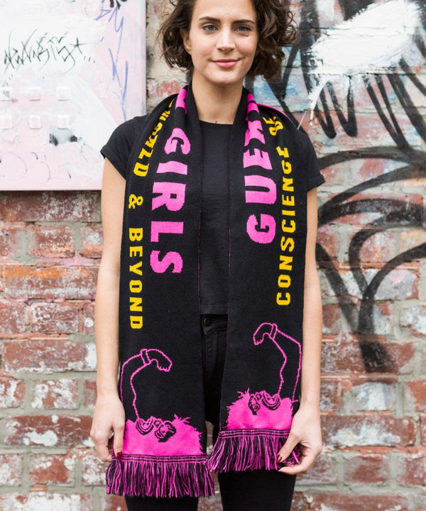 A woman wearing a scarf standing before a brick wall. The pink and black scarf is in the style of a soccer scarf with an illustration of a person in a gorilla mask holding up their fist and text in pink and yellow.