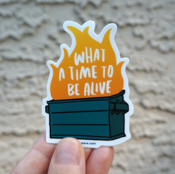 A concrete background with a hand holding a sticker before it. The sticker featured is a dumpster with a fire in it. There are words in the fire that read "What a Time to be Alive."