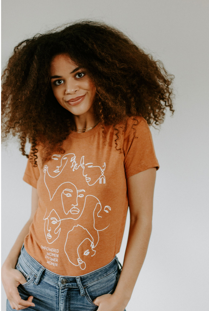 Woman with a dark skin tone wearing an orange t-shirt with white line illustrations of women's faces.