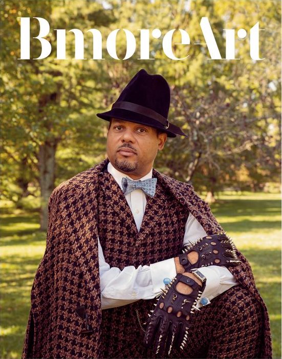 A magazine cover with a photograph of a man with a dark skin tone in tweed and a felt hat sitting before a forest. The title reads "Bmore Art."