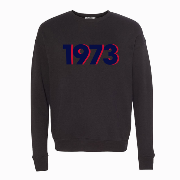 A crewneck in a washed-out vintage black color with the letters "1973" printed on it in blue and red.