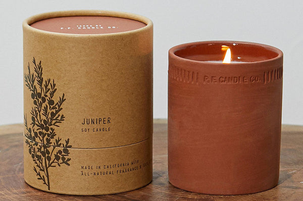A round paper box with the text "Juniper: Soy Candle" next to a candle in a red clay cup, burning.