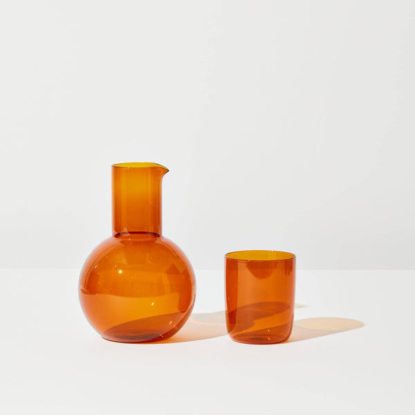 A glass carafe and cup in an amber color standing before a white background.