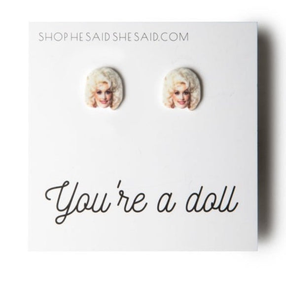 Two stud earrings with little faces of a woman with big blonde hair. The text underneath on the paper reads "You're a doll."