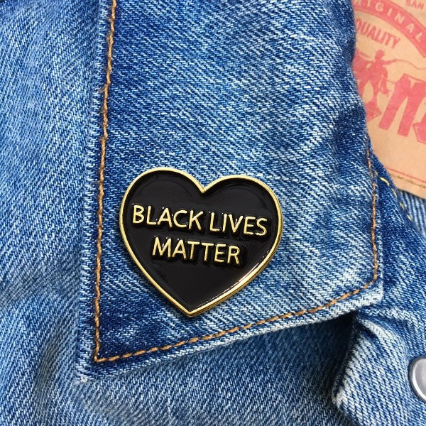 A black enamel pin in the shape of a heart with golden lettering. The text reads "Black Lives Matter" in all caps. The pin is attached to a denim jacket.