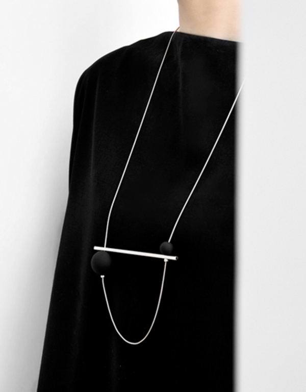 A woman wearing a necklace, a mixed media designed geometric necklace featuring a black bar and two white orbs.