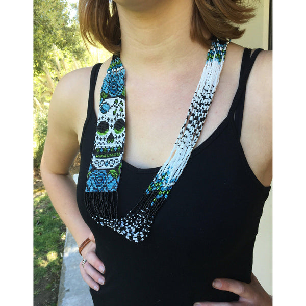 A woman with a light skin tone is wearing a beaded necklace in black, white, blue, and green and with a skull pattern.