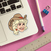 A sticker of a woman's head on a lapotp. The woman has a light skin tone, blonde hair, and is wearing a cowboy hat. On her hat, it says "Dolly."