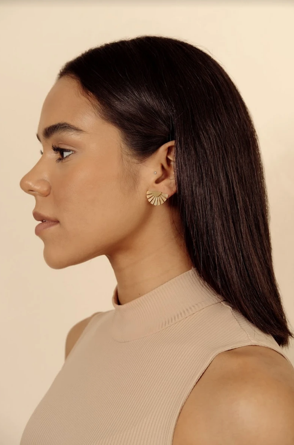 A woman with a medium-dark skin tone is wearing a brass half moon stud earring in gold. The earring is dainty and resembles a sun.