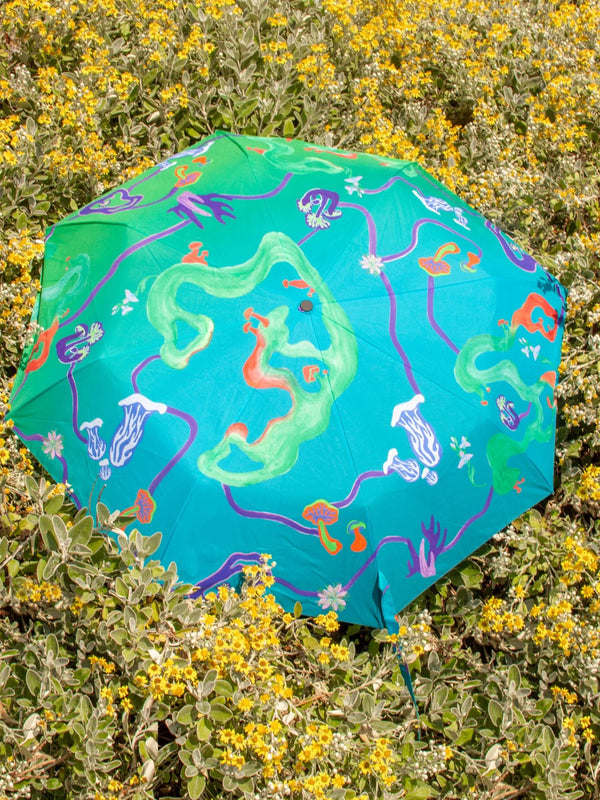 An opened up umbrella lying on a bed of flowers. The umbrella is in a teal color and features mushrooms in lilac and blue.