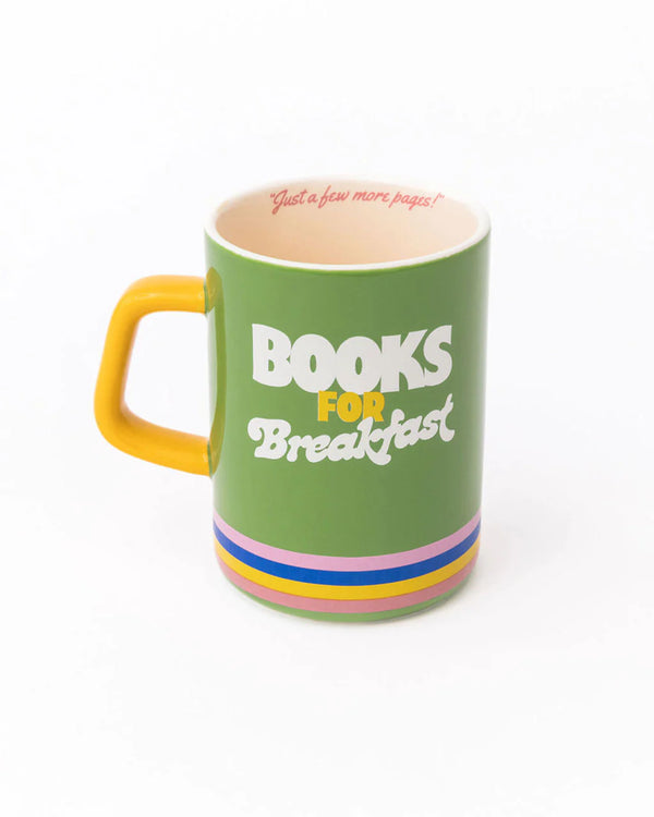 A white background with a green mug before it . The mug has four bands on the bottom in the colors of pink, blue, yellow, and coral. The handle is yellow and the interior of the cup is beige with the words "Just a Few More Pages!" written inside in pink cursive. On the front of the cup are the words "Books For Breakfast" Written in bold font. 